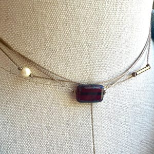 May include: Delicate layered necklace featuring a rectangular, dark red and blue glass bead. The necklace also includes a small pearl and a gold-colored bar. The necklace is displayed on a neutral background.