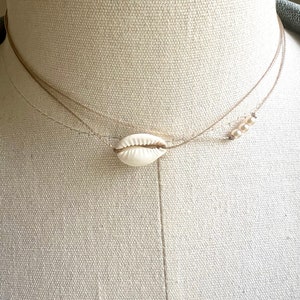 May include: A delicate layered necklace with a white seashell pendant and small white beads. The necklace is made of thin brown cord and is displayed on a white mannequin.