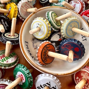 May include: A collection of colorful bottle caps with wooden dowels inserted through the center. The bottle caps have various designs and text, including "Orange Soda", "Stay Scky", "Decaffeinated Coffee", and "Ginger Ale".