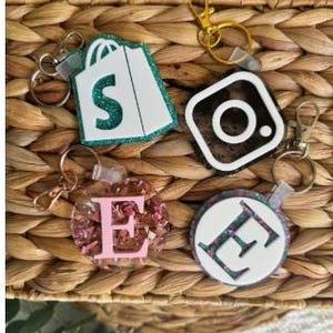 May include: A collection of keychains with various designs. One keychain features a white shopping bag with a green glitter outline and the letter "S". Other keychains include an Instagram logo, a pink floral "E", and a white circle with a glitter "E".