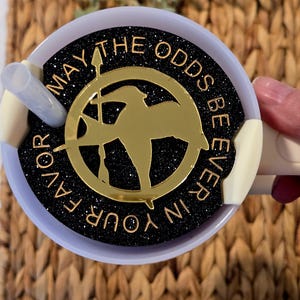 Puede incluir: Una tapa de taza de purpurina negra y dorada con el texto "Que la suerte est&eacute; siempre de tu lado" y un pin de sinsajo dorado.