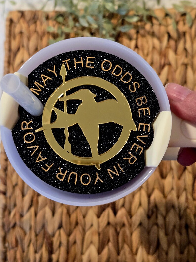 Puede incluir: Una tapa de taza de purpurina negra y dorada con el texto "Que la suerte est&eacute; siempre de tu lado" y un pin de sinsajo dorado.