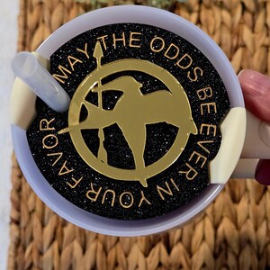 Puede incluir: Un vaso de glitter negro y dorado con una silueta dorada de sinsajo y el texto "Que la suerte est&eacute; siempre de tu lado".