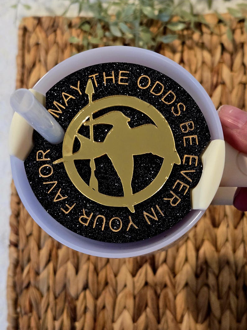 Puede incluir: Un vaso de glitter negro y dorado con una silueta dorada de sinsajo y el texto "Que la suerte est&eacute; siempre de tu lado".