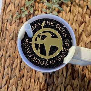 Puede incluir: Un vaso negro y dorado con acabado brillante y un dise&ntilde;o de metal dorado con un sinsajo y el texto "Que la suerte est&eacute; siempre de tu lado".