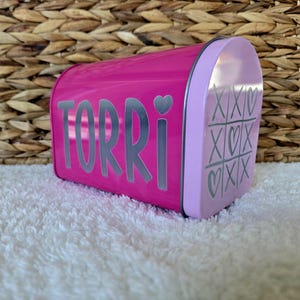 May include: A pink metal tin with the name "TORRI" in silver letters on the front. The side of the tin has a pink and white design with hearts and X's.