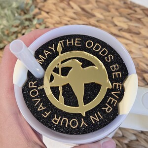 Puede incluir: Un vaso blanco con una tapa negra y un dise&ntilde;o de purpurina dorada. La tapa tiene un pin dorado de sinsajo con las palabras "Que la suerte est&eacute; siempre de tu lado".