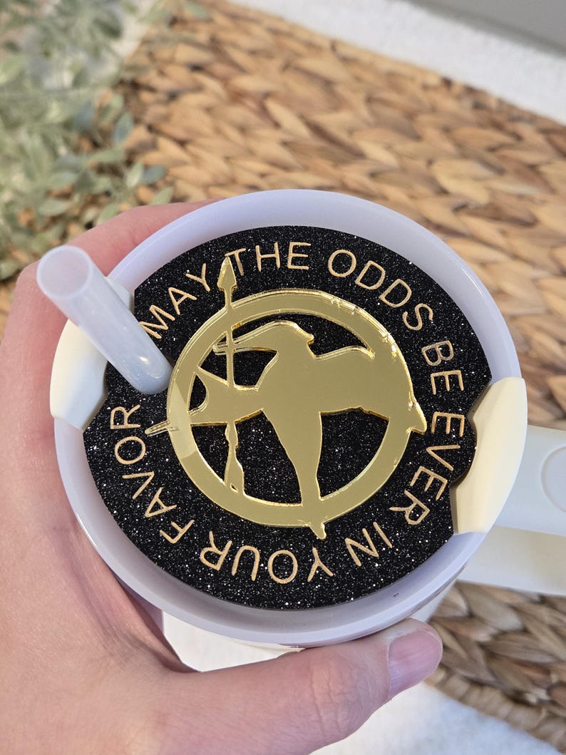 Puede incluir: Un vaso blanco con una tapa negra y un dise&ntilde;o de purpurina dorada. La tapa tiene un pin dorado de sinsajo con las palabras "Que la suerte est&eacute; siempre de tu lado".