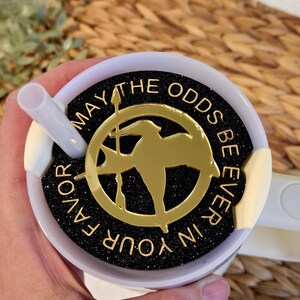 Puede incluir: Una taza de viaje blanca con una tapa negra y dorada. La tapa tiene un fondo negro con purpurina dorada y un pin dorado de sinsajo con las palabras "Que la suerte est&eacute; siempre de tu lado".