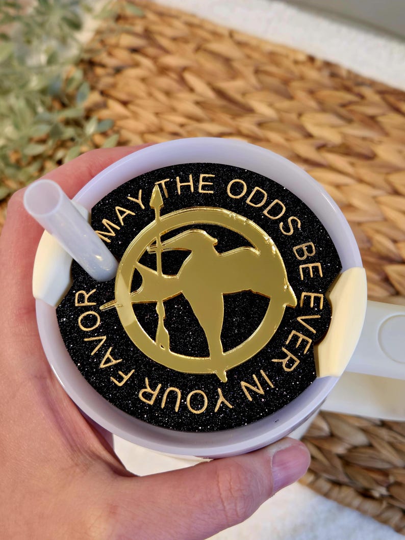 Puede incluir: Una taza de viaje blanca con una tapa negra y dorada. La tapa tiene un fondo negro con purpurina dorada y un pin dorado de sinsajo con las palabras "Que la suerte est&eacute; siempre de tu lado".