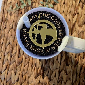 Puede incluir: Un vaso de glitter negro y dorado con tapa blanca y una pajita. El vaso presenta un pin de sinsajo dorado con el texto "Que la suerte est&eacute; siempre de tu lado".
