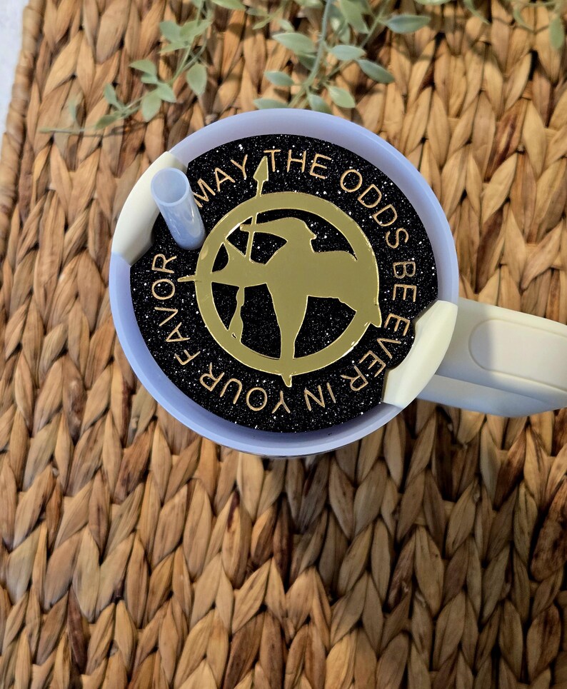 Puede incluir: Un vaso de glitter negro y dorado con tapa blanca y una pajita. El vaso presenta un pin de sinsajo dorado con el texto "Que la suerte est&eacute; siempre de tu lado".