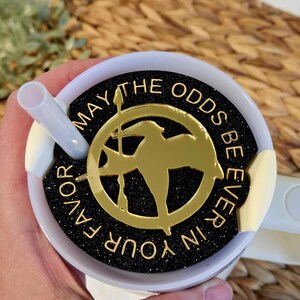 Puede incluir: Taza de viaje blanca con una tapa negra y dorada. La tapa tiene un fondo negro con purpurina dorada y las palabras "Que la suerte est&eacute; siempre de tu lado" escritas en dorado. Un pin dorado de sinsajo est&aacute; en el centro de la tapa.
