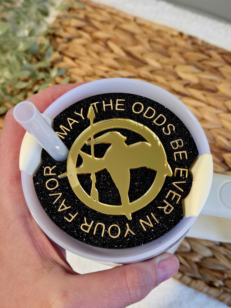 Puede incluir: Taza de viaje blanca con una tapa negra y dorada. La tapa tiene un fondo negro con purpurina dorada y las palabras "Que la suerte est&eacute; siempre de tu lado" escritas en dorado. Un pin dorado de sinsajo est&aacute; en el centro de la tapa.
