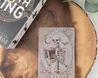 Puede incluir: Una carta de tarot blanca con una ilustración en blanco y negro de un esqueleto leyendo un libro. La carta tiene un borde decorativo y el texto "THE READER" en la parte inferior.