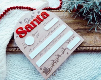 Puede incluir: Cartel de Navidad de madera con las palabras "Querido Papá Noel" en letras 3D rojas. El cartel tiene espacios para escribir información sobre la edad del niño, su cosa favorita de la Navidad y su deseo navideño. Un trineo de renos está grabado en la parte inferior.