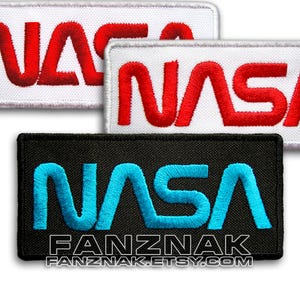 May include: Three embroidered NASA patches. One patch is white with red lettering, one is white with red lettering, and one is black with blue lettering. The patches are all rectangular and have a stitched border.