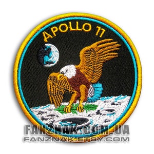 May include: A round, yellow-bordered patch with a black background. The patch features a white eagle with brown feathers landing on a white surface with grey craters. The eagle has a blue wing and a white head. The text "APOLLO 11" is in yellow above the eagle. A blue and white globe is in the top left corner.