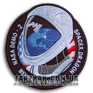 May include: A black and white embroidered patch featuring a white astronaut helmet with a blue and green Earth inside. The text "NASA DEMO-2 SPACEX DRAGON" is embroidered around the patch.