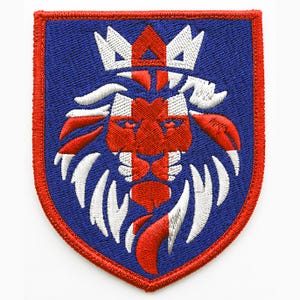 May include: A shield-shaped embroidered patch featuring a stylised lion head design in red and white thread against a solid blue background. The lion is depicted with a crown and a flowing mane, all enclosed by a thick, stitched red border.