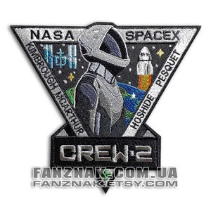 May include: A black and white embroidered patch with a triangular shape. The patch features a stylised astronaut in a spacesuit, a rocket, and the text "NASA SPACEX CREW-2" with the names of the astronauts "KIMBROUGH, McARTHUR, HOSHIDE, PESQUET" around the perimeter.