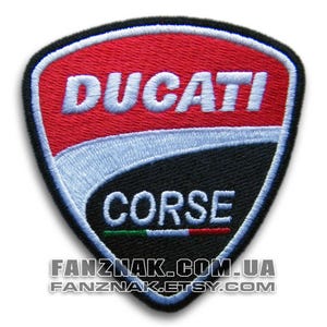 May include: Embroidered patch with the Ducati Corse logo. The shield-shaped patch features a red top, a white and light blue central section, and a black base. The words "DUCATI" and "CORSE" are embroidered in white.
