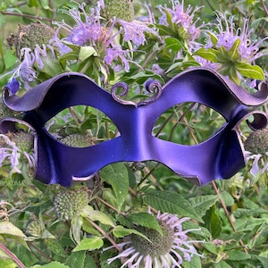 May include: A deep purple masquerade mask with a metallic sheen, featuring curved edges and decorative swirls above the eye openings. The mask is set against a backdrop of green foliage and purple flowers, creating a striking contrast.