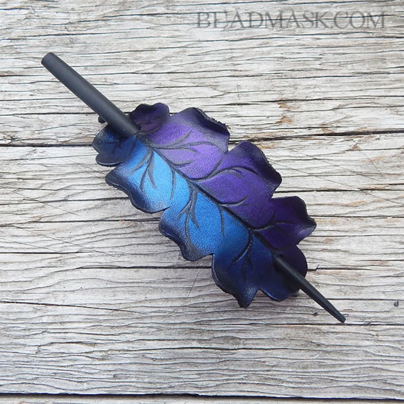 May include: A handcrafted hair barrette featuring a leaf design with a gradient of blue and purple hues. The barrette has a dark gray stick for securing the hair. The website beadmask.com is visible in the image.