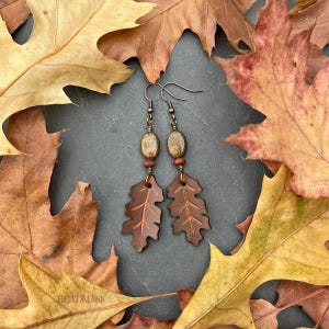 Peut inclure: Boucles d'oreilles artisanales avec des pendentifs en forme de feuilles de ch&ecirc;ne en cuir marron, des perles ovales et des d&eacute;tails cuivr&eacute;s. Les boucles d'oreilles sont pr&eacute;sent&eacute;es sur un fond de feuilles d'automne.