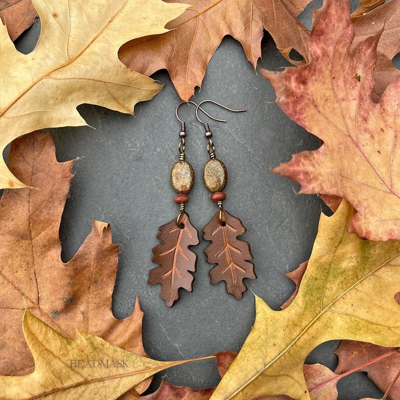 Peut inclure: Boucles d'oreilles artisanales avec des pendentifs en forme de feuilles de ch&ecirc;ne en cuir marron, des perles ovales et des d&eacute;tails cuivr&eacute;s. Les boucles d'oreilles sont pr&eacute;sent&eacute;es sur un fond de feuilles d'automne.