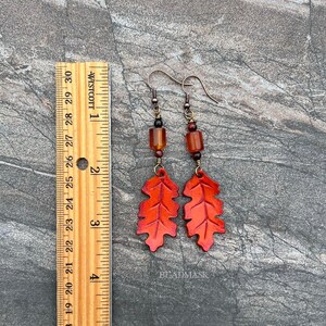 Peut inclure: Boucles d'oreilles faites &agrave; la main avec des pendentifs en forme de feuilles orange vif. Les boucles d'oreilles ont des crochets en m&eacute;tal fonc&eacute; et des perles marron. Les feuilles mesurent environ 5 cm de long. Le mot "BEADMASK" est visible.