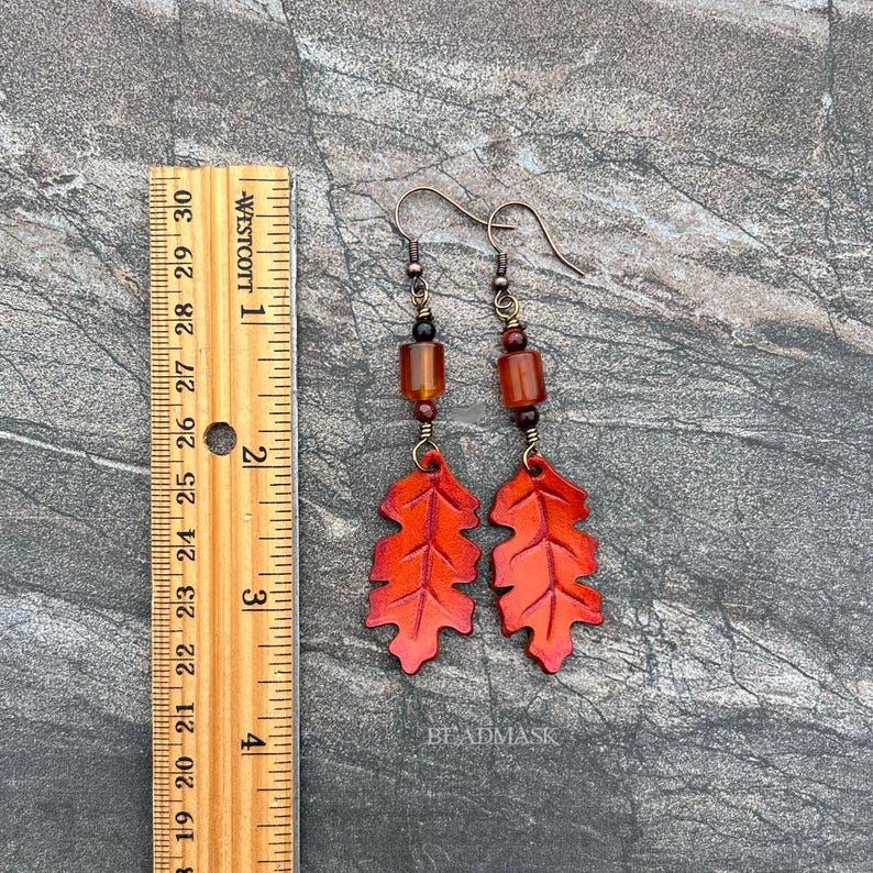Peut inclure: Boucles d'oreilles faites &agrave; la main avec des pendentifs en forme de feuilles orange vif. Les boucles d'oreilles ont des crochets en m&eacute;tal fonc&eacute; et des perles marron. Les feuilles mesurent environ 5 cm de long. Le mot "BEADMASK" est visible.