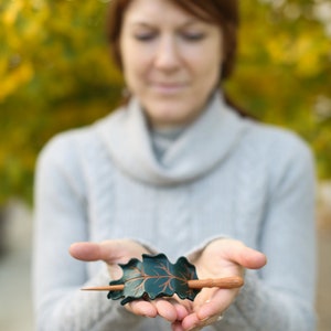 May include: A handcrafted hair barrette featuring a dark green oak leaf design with copper-colored accents. The barrette includes a wooden stick for securing the hair. The accessory is held in open hands.