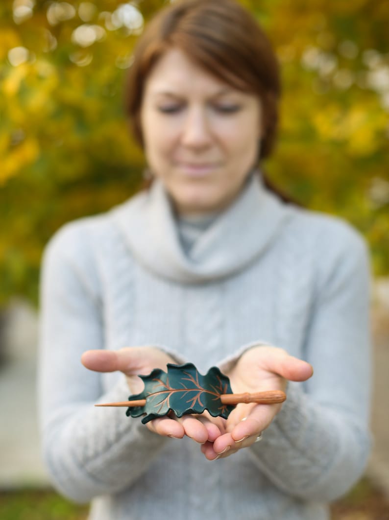 May include: A handcrafted hair barrette featuring a dark green oak leaf design with copper-colored accents. The barrette includes a wooden stick for securing the hair. The accessory is held in open hands.