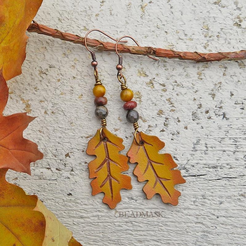 Peut inclure: Boucles d'oreilles artisanales avec des pendentifs en forme de feuilles jaunes et oranges, avec des d&eacute;tails de veines complexes. Les boucles d'oreilles sont orn&eacute;es d'une s&eacute;rie de perles color&eacute;es et de fil de cuivre. Elles sont pr&eacute;sent&eacute;es sur une surface en bois.