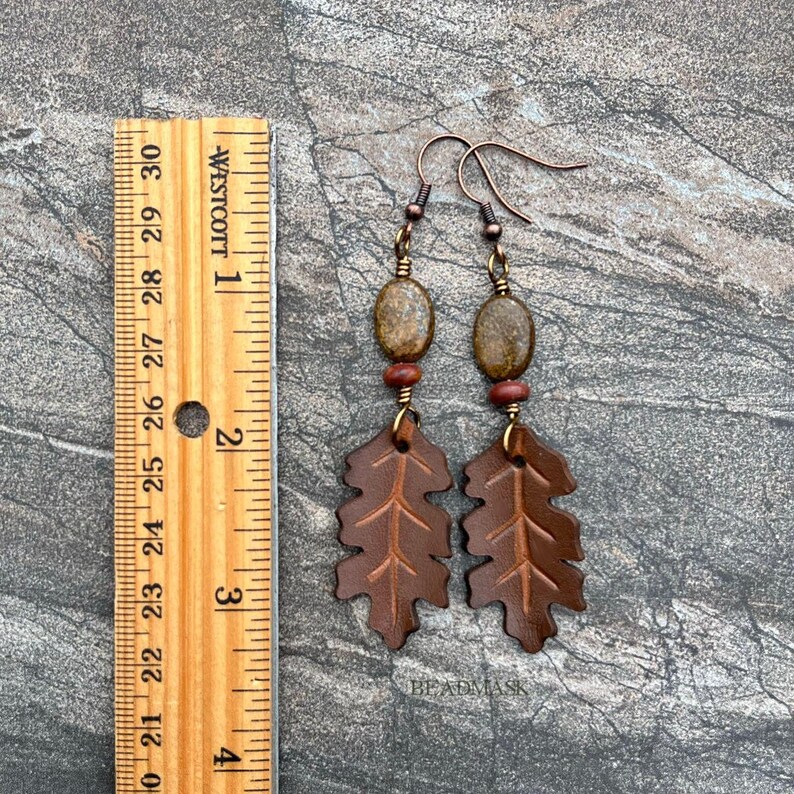 Peut inclure: Boucles d'oreilles artisanales avec des pendentifs en forme de feuilles de ch&ecirc;ne en cuir marron, des perles ovales et des crochets d'oreilles cuivr&eacute;s. Mesurent environ 7.6 cm de long, avec un design rustique inspir&eacute; de la nature.