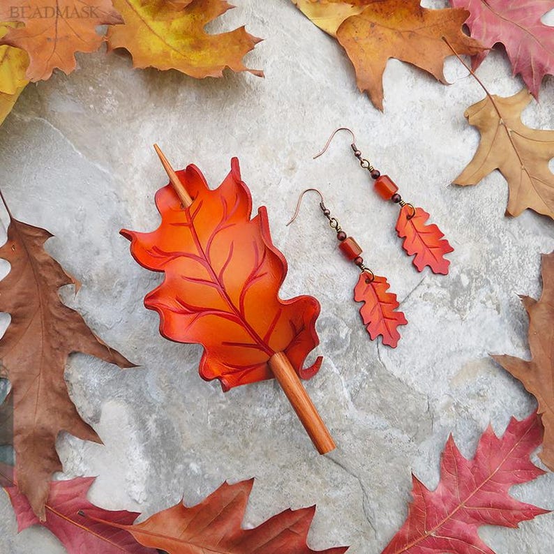 Peut inclure: Un ensemble de bijoux sur le th&egrave;me de l'automne. L'image pr&eacute;sente une barrette &agrave; cheveux en forme de feuille orange avec un manche en bois, et une paire de boucles d'oreilles avec des pendentifs en forme de feuille et des perles. Le tout est pr&eacute;sent&eacute; avec des feuilles mortes.