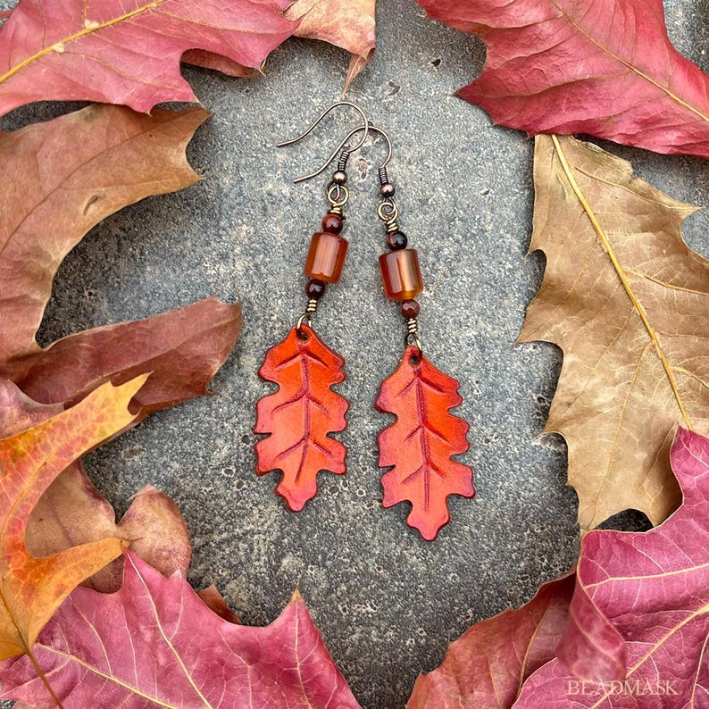 Peut inclure: Boucles d'oreilles artisanales sur le th&egrave;me de l'automne. Elles pr&eacute;sentent un motif de feuille d'un rouge-orang&eacute; vif. Elles sont orn&eacute;es de perles marron et de crochets en m&eacute;tal fonc&eacute;, sur un fond de feuilles d'automne.