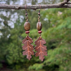 Peut inclure: Boucles d'oreilles artisanales avec des pendentifs en forme de feuilles de cuir marron. Chaque boucle d'oreille comporte une perle ovale marron fonc&eacute; et de petites perles rouges. Elles sont suspendues &agrave; des crochets d'oreilles cuivr&eacute;s, sur fond vert flou.