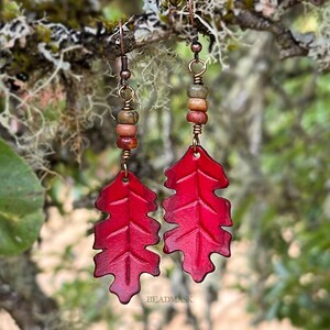 Peut inclure: Une paire de boucles d'oreilles rouges en forme de feuille avec des fils et des perles de couleur cuivre. Les boucles d'oreilles sont suspendues &agrave; des crochets d'oreilles couleur cuivre. Le fond est une vue floue d'une branche d'arbre et de feuillage. Le texte "BEADMASK" est visible.