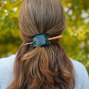 May include: A dark green leaf-shaped hair barrette with a copper-colored vein design and a wooden stick closure. The barrette is set in brown hair, with a blurred green and yellow background.