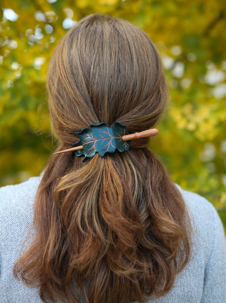 May include: A dark green leaf-shaped hair barrette with a copper-colored vein design and a wooden stick closure. The barrette is set in brown hair, with a blurred green and yellow background.