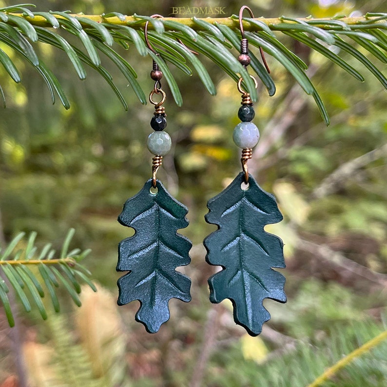 Peut inclure: Boucles d'oreilles en forme de feuilles en cuir vert fonc&eacute;, avec des crochets et des accents de fil cuivr&eacute;s. Chaque boucle d'oreille pr&eacute;sente une forme de feuille d&eacute;taill&eacute;e, avec de petites perles vertes et noires. Les boucles d'oreilles sont suspendues &agrave; une branche verte.