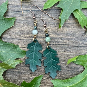 Peut inclure: Boucles d'oreilles artisanales avec des pendentifs en forme de feuilles de ch&ecirc;ne en cuir vert fonc&eacute;. Les boucles d'oreilles ont des crochets d'oreille cuivr&eacute;s et sont orn&eacute;es de perles de pierres pr&eacute;cieuses vertes et grises. Fond avec des feuilles de ch&ecirc;ne vertes.