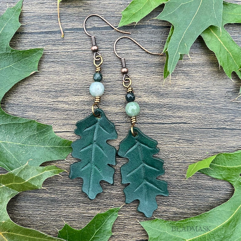 Peut inclure: Boucles d'oreilles artisanales avec des pendentifs en forme de feuilles de ch&ecirc;ne en cuir vert fonc&eacute;. Les boucles d'oreilles ont des crochets d'oreille cuivr&eacute;s et sont orn&eacute;es de perles de pierres pr&eacute;cieuses vertes et grises. Fond avec des feuilles de ch&ecirc;ne vertes.