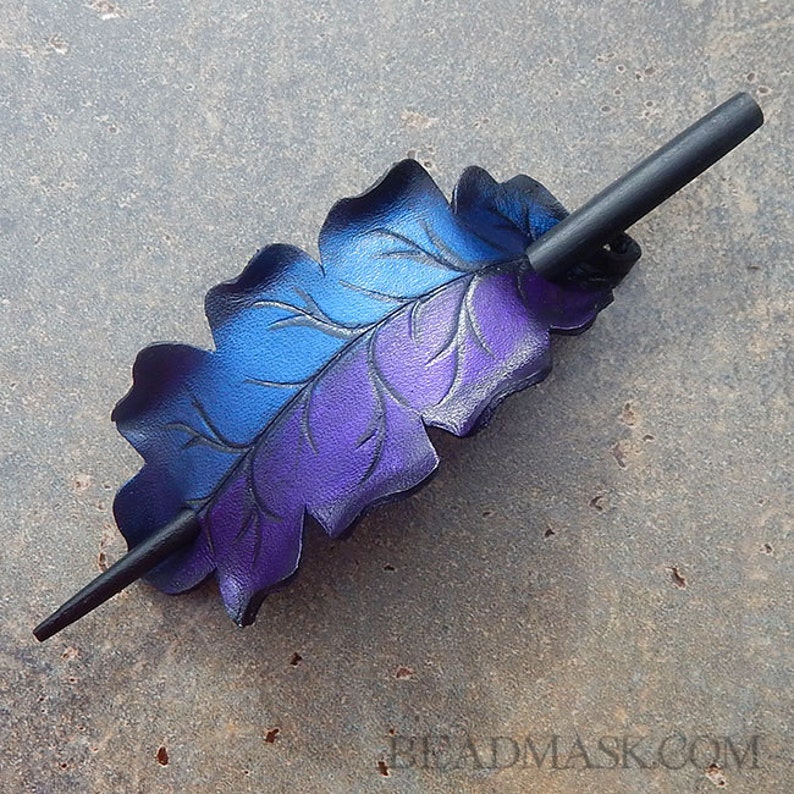May include: A handcrafted hair barrette featuring a leaf design with blue and purple gradient coloring. The barrette includes a dark wooden stick for securing the hair accessory. The leaf has detailed vein patterns.