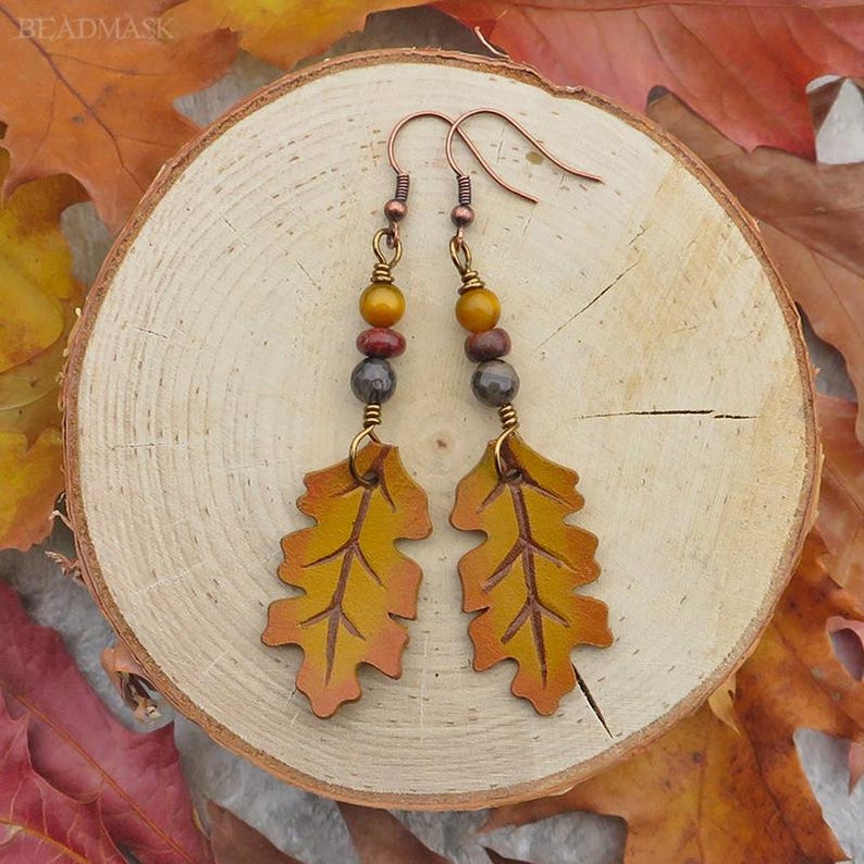 Peut inclure: Boucles d'oreilles artisanales avec des pendentifs en forme de feuilles aux tons jaune et orange, avec des d&eacute;tails de veines complexes. Les boucles d'oreilles ont un accent de perles jaunes, rouges et grises, et des crochets d'oreilles cuivr&eacute;s. Elles sont pr&eacute;sent&eacute;es sur une tranche de bois.
