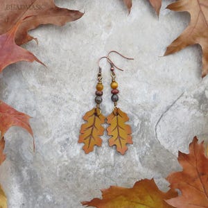 Peut inclure: Une paire de boucles d'oreilles sur le th&egrave;me de l'automne. Chaque boucle d'oreille pr&eacute;sente un pendentif en forme de feuille de ch&ecirc;ne de couleur cuivre avec des nuances de jaune et d'orange. Les boucles d'oreilles ont un accent de perles marron, jaunes et dor&eacute;es. Le fond est une surface de pierre gris clair avec des feuilles d'automne &eacute;parpill&eacute;es.