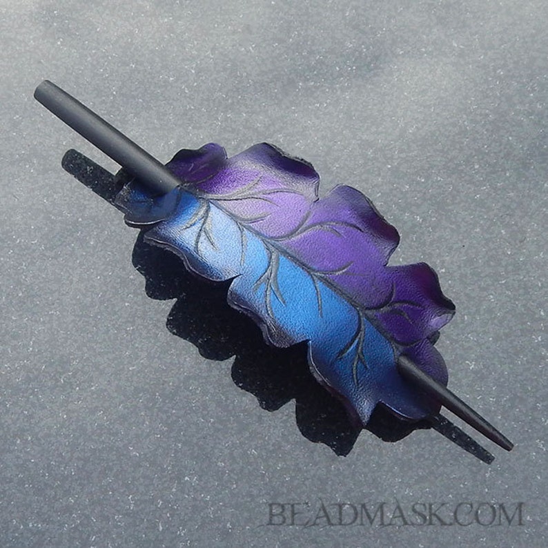 May include: A handcrafted hair accessory featuring a leaf design with a gradient of blue and purple hues. The leaf is detailed with etched veins and is paired with a black stick. The accessory is a unique hair piece.