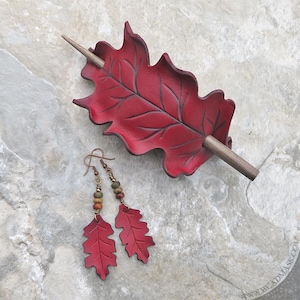 Peut inclure: Un barrette &agrave; cheveux en forme de feuille de cuir rouge avec un b&acirc;ton en bois, et une paire de boucles d'oreilles en forme de feuille assorties. Les boucles d'oreilles ont des crochets couleur cuivre et des accents perl&eacute;s verts, bruns et dor&eacute;s. Sur une surface en pierre.